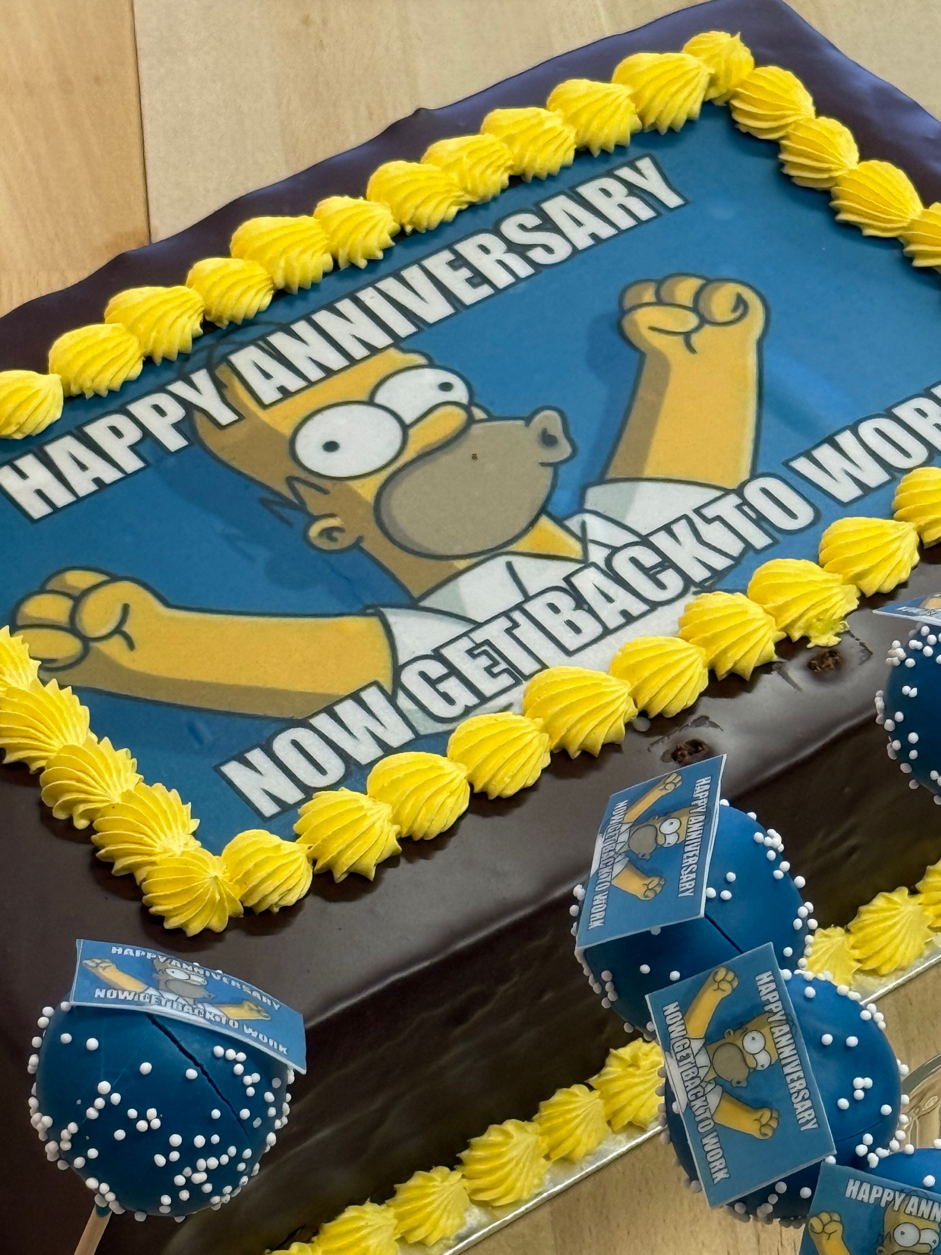 Rechteckige Schokoladentorte mit gelber Verzierung und Aufschrift „Happy Anniversary, Now Get Back to Work“ auf einer bedruckten Zuckerfolie mit einer bekannten Cartoon-Figur.