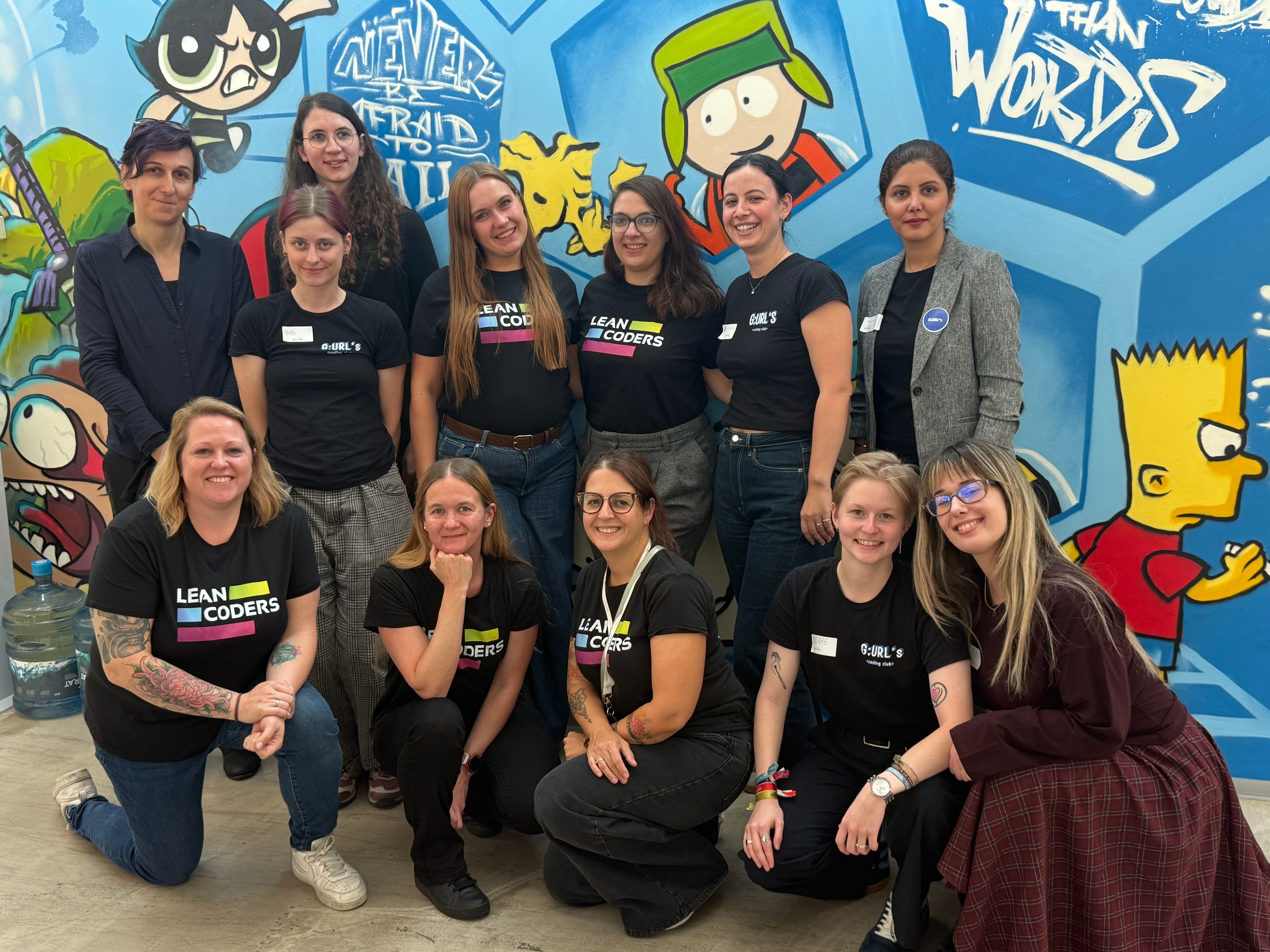 Gruppenfoto von zehn Frauen, die in schwarzen T-Shirts mit dem Aufdruck „LEAN CODERS“ vor einer graffitiartigen Wand stehen. Die Hintergründe zeigen Charaktere aus Cartoons. Sie lächeln und posieren zusammen, während einige mit Namensschildern ausgestattet sind.