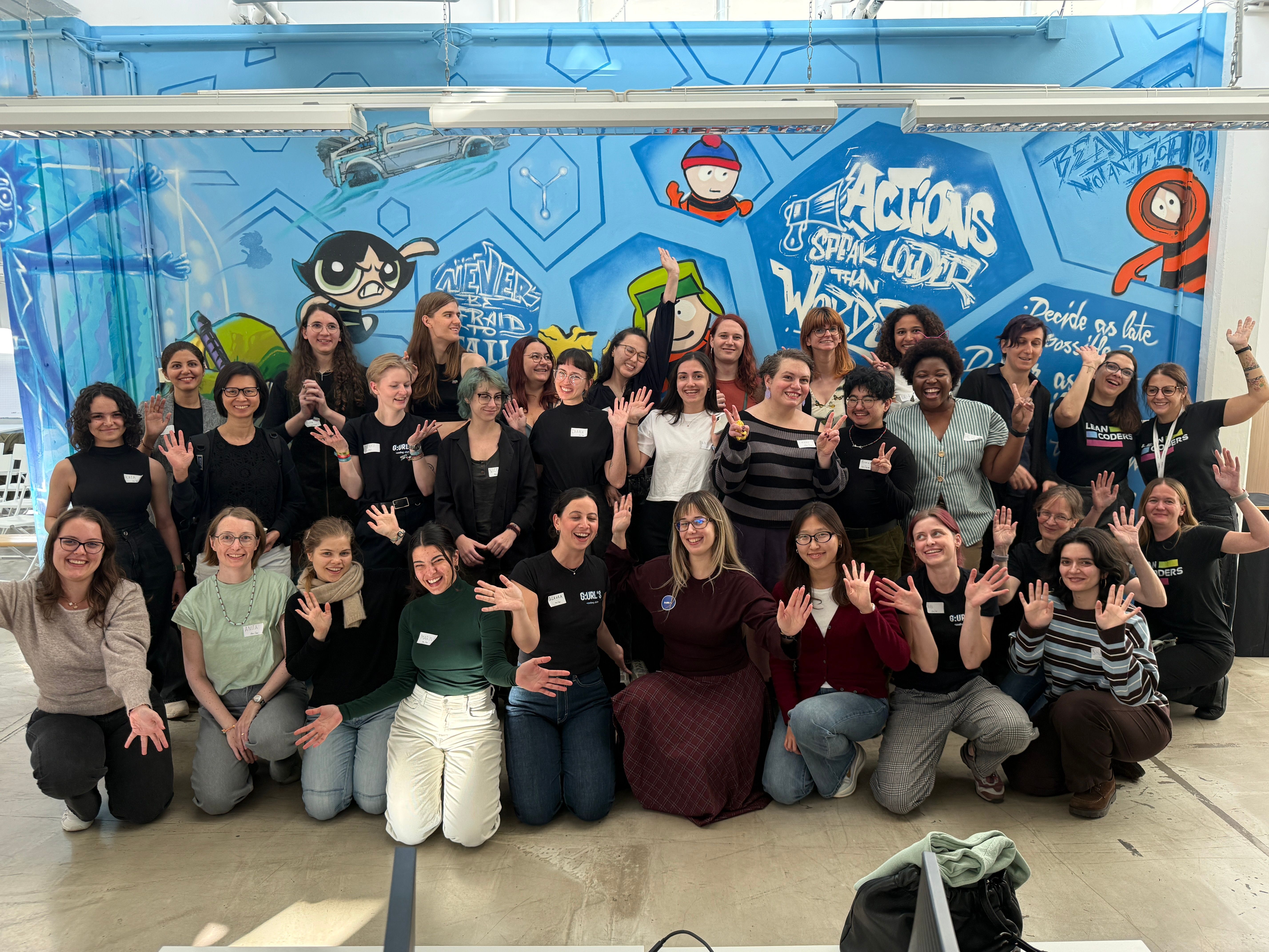 Gruppenfoto von 38 Frauen, die sich vor einer bunten Wand mit Graffiti versammeln und lächeln. Viele Teilnehmerinnen haben die Hände gehoben, um zu winken oder Zeichen zu machen. Die Wand zeigt verschiedene Charaktere und inspirierende Sprüche, die die Atmosphäre von Gemeinschaft und Unterstützung vermitteln.