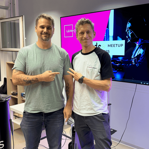 Zwei Männer stehen in einem modernen Büro und posieren entspannt für die Kamera. Im Hintergrund ist ein Bildschirm mit der Aufschrift „LEAN-CODERS MeetUp“ und einem grafischen Element zu sehen. Der Mann links trägt ein graues T-Shirt und hat kurze, helle Haare, während der Mann rechts ein schwarzes und weißes T-Shirt trägt und lockige, hellbraune Haare hat.