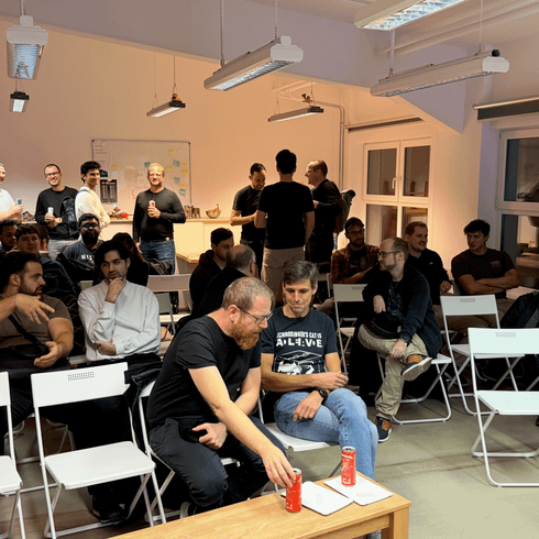 Gruppenszene in einem Veranstaltungsraum mit mehreren Personen, die am Tisch sitzen, Bier trinken und miteinander sprechen. Im Hintergrund stehen weitere Teilnehmer und interagieren. Die Stühle sind weiß und die Wände sind hell, mit Tisch und Notizen auf einer Wandtafel sichtbar.