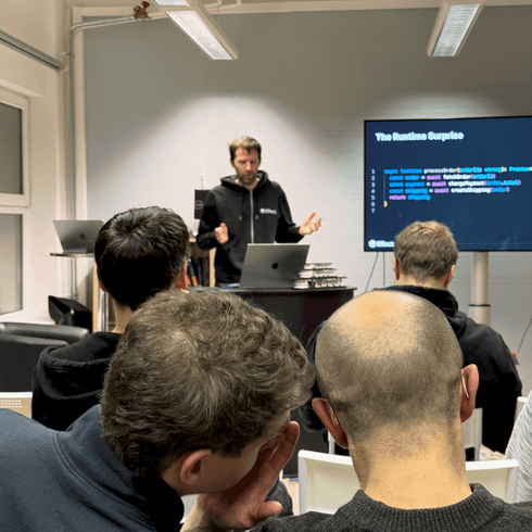 Junger Mann in schwarzem Hoodie präsentiert vor einer Gruppe von Zuhörern, die ihre Aufmerksamkeit auf den Bildschirm richten, auf dem Programmiercode angezeigt wird. Drei Personen im Vordergrund sind zu sehen, während sie sich unterhalten.