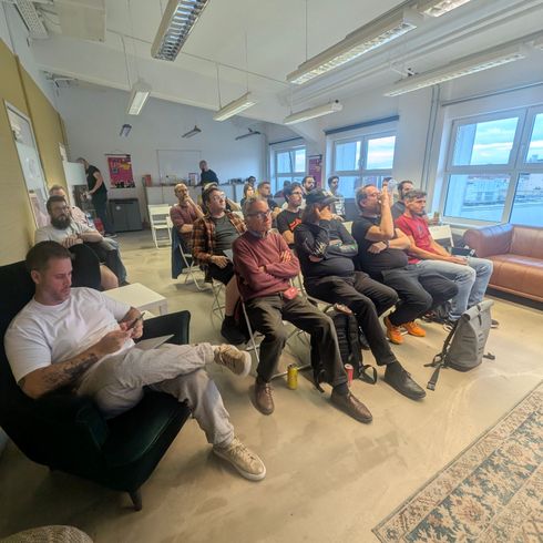 Eine Gruppe von Personen sitzt in einem modernen Raum, einige hören aufmerksam zu, während andere mit ihren Mobiltelefonen beschäftigt sind. Im Vordergrund sitzt ein junger Mann in heller Kleidung auf einem bequemen Sessel und schaut auf sein Gerät. Im Hintergrund sind Tische und Fenster sichtbar, die einen Blick nach draußen bieten.