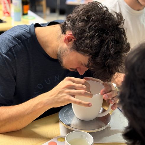 Ein Mann bemalt konzentriert eine weiße Tasse, während er hellfarbige Farben und Wasser auf einem Holztisch nutzt.