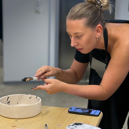 Eine Frau bemalt mit einem Pinsel eine weiße Tonschale auf einem Tisch, umgeben von Malutensilien.