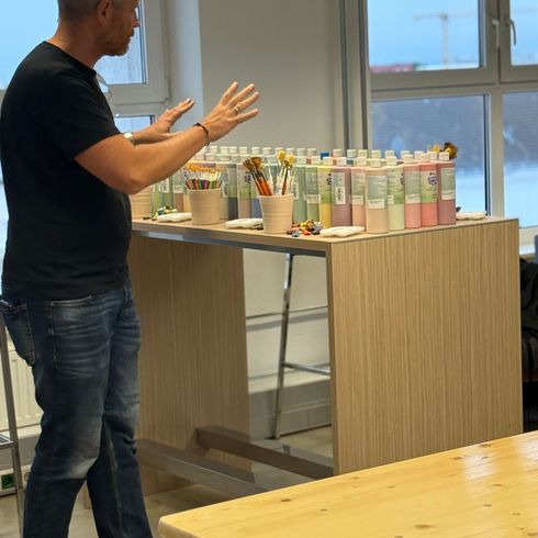Person erklärt einem kleinen Publikum Malzubehör, darunter Pinsel und Farben, auf einem Tisch in einem offenen, hell beleuchteten Raum.