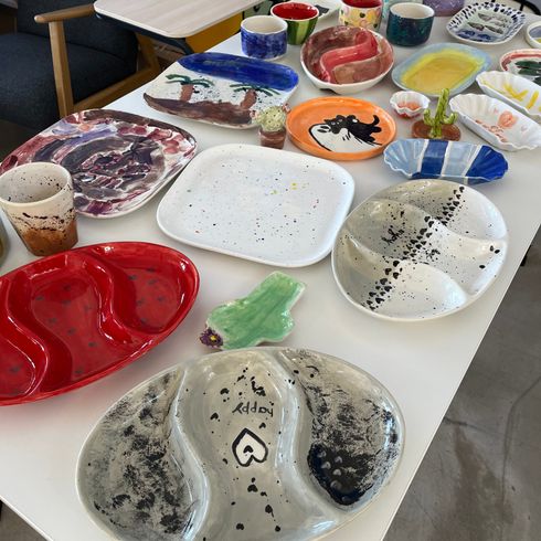 Verschiedenfarbig bemalte Keramikschalen und Tassen auf einem Tisch, mit einem farbenfrohen Wandgemälde im Hintergrund.