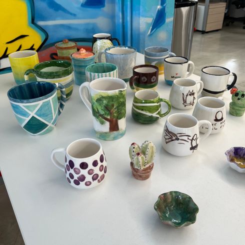 Verschiedene farbige und gemusterte Keramikbecher und kleine Schalen auf einem weißen Tisch vor einer bunt bemalten Wand.