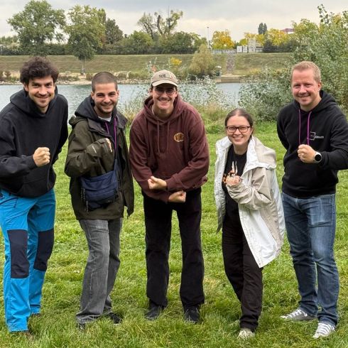 Fünf Personen stehen lächelnd in einer grünen Wiese vor einem Fluss, alle zeigen eine entschlossene Pose mit geballten Fäusten.