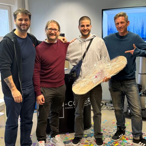 Vier Männer stehen lächelnd in einem Büro, einer von ihnen hält ein großes, in Folie verpacktes Geschenk. Auf dem Boden liegen bunte Papierkonfetti.