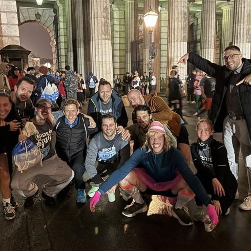 Gruppe von Menschen in sportlicher Kleidung posiert gut gelaunt vor einem erleuchteten Tor mit zahlreichen weiteren Personen im Hintergrund.