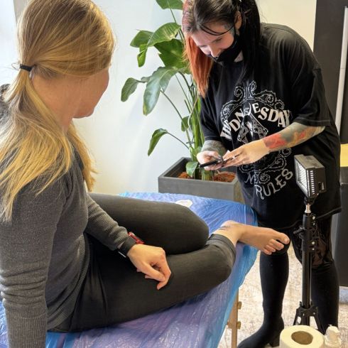 Eine Frau sitzt auf einer Liege, während eine Tätowiererin ein Tattoo auf ihren Fuß vorbereitet. Im Hintergrund ist eine Pflanze zu sehen.