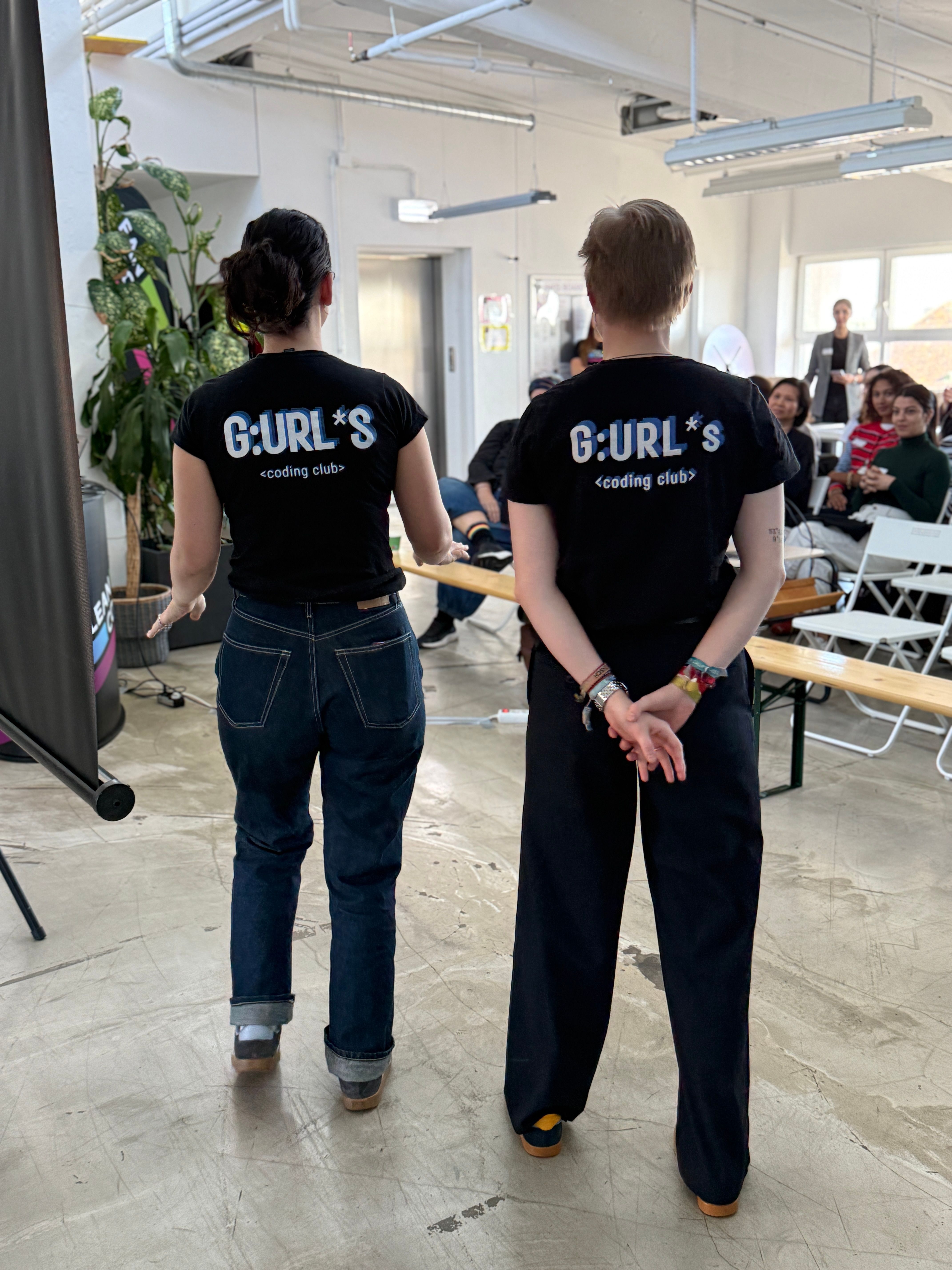 Zwei Personen stehen Rücken an Rücken, beide tragen schwarze T-Shirts mit der Aufschrift „GURL'S <coding club>“. Im Hintergrund ist eine Gruppe von Menschen in einem Seminarraum zu sehen, mit Sitzgelegenheiten und Pflanzen.