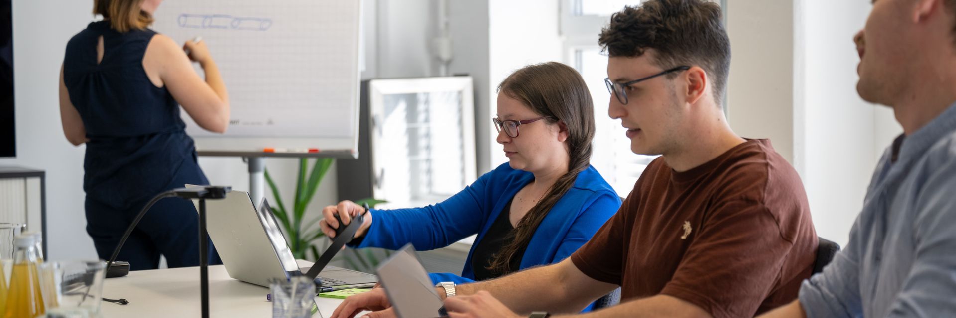 Personen in einem Besprechungsraum, die an Laptops arbeiten, während eine Frau an einem Flipchart zeichnet.