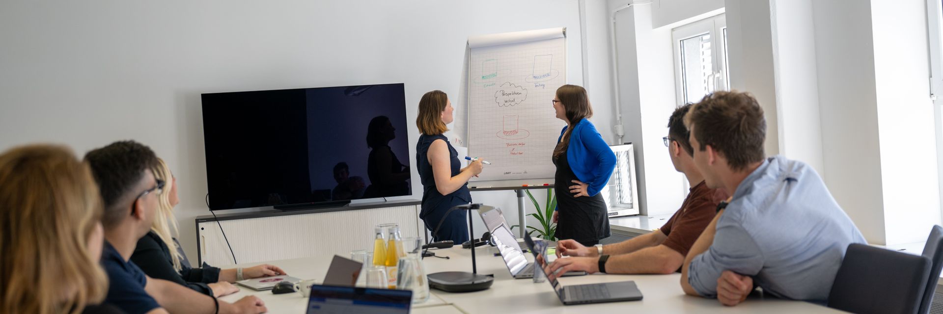 Personengruppe bei einem Meeting in einem Konferenzraum, eine Frau präsentiert an einem Whiteboard, während andere Teilnehmer aufmerksam zuhören und Notizen machen.