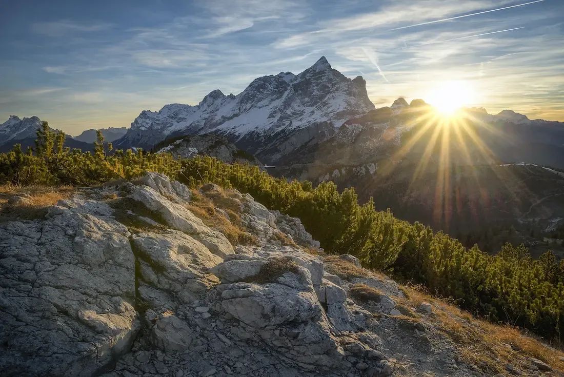 Sonnenaufgang über einem schneebedeckten Bergmassiv mit felsigem Vordergrund und grünen Kiefern im Vordergrund.