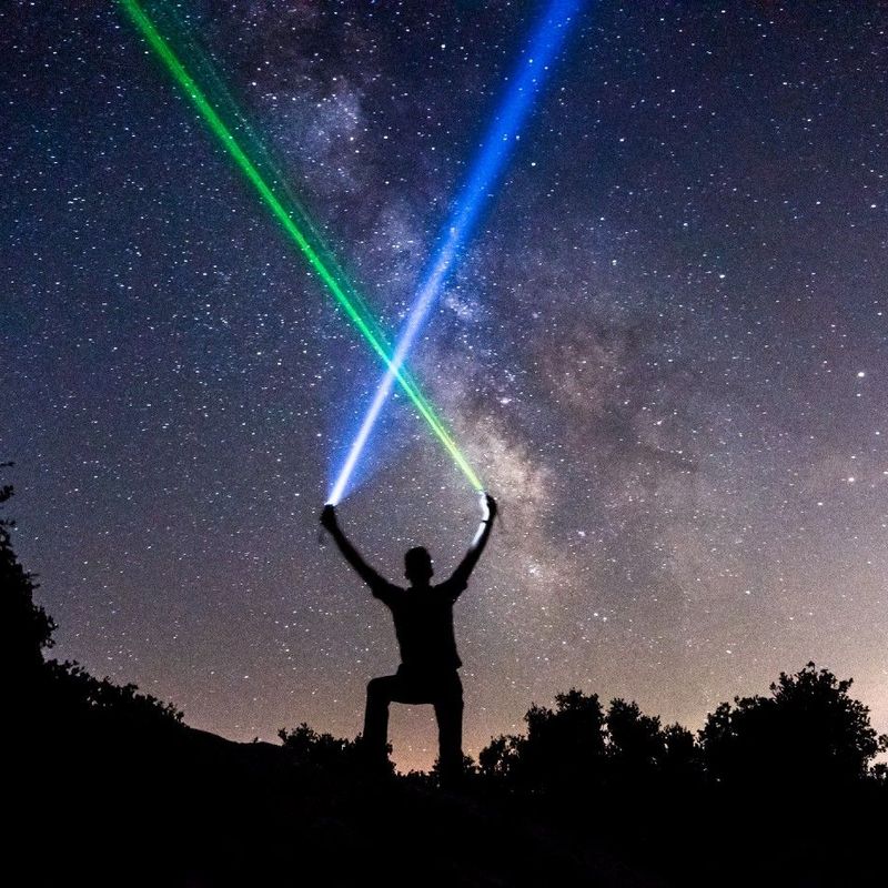 Person hält zwei Laser unter sternklarem Himmel, mit der Milchstraße im Hintergrund.