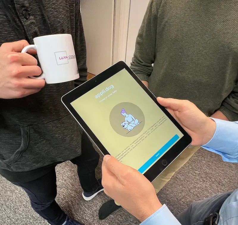Person hält ein Tablet mit einer geöffneten App, während zwei andere Personen daneben stehen; eine hält eine Kaffeetasse.