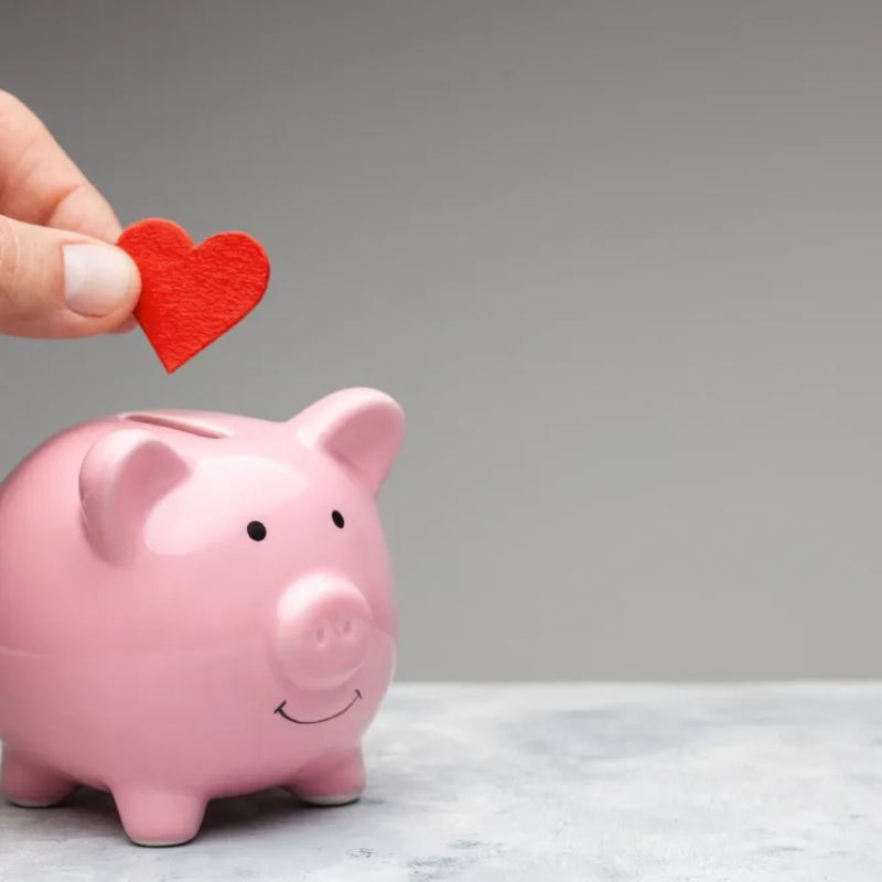 Hand legt ein rotes Herz in ein rosa Sparschwein.