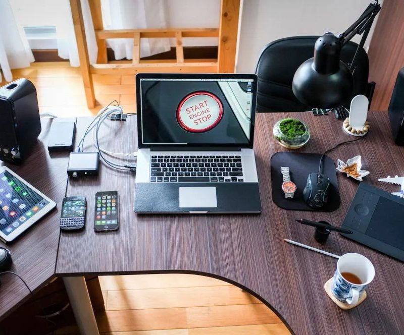 Moderner Schreibtisch mit verschiedenen Elektronikgeräten, darunter ein Laptop, mehrere Smartphones, ein Tablet und Kopfhörer, sowie Bürozubehör wie eine Schreibtischlampe und eine Tasse Tee.
