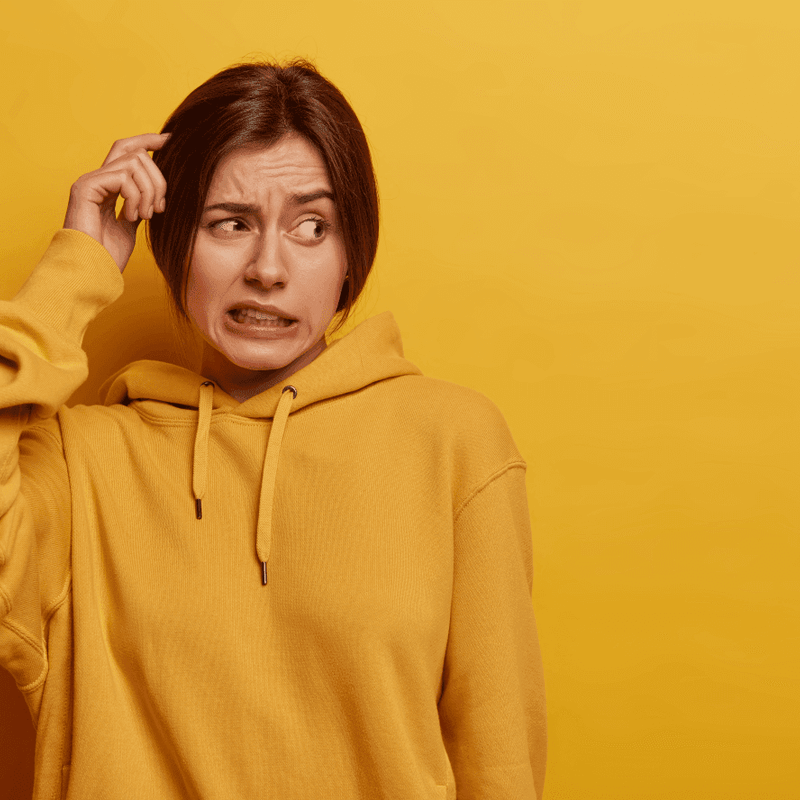 Person in einem gelben Hoodie schaut verwirrt und kratzt sich am Kopf, vor einem einfarbig gelben Hintergrund.