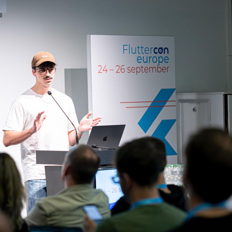 Redner gibt eine Präsentation auf der Fluttercon Europe vor einem Publikum, neben einem Schild mit dem Datum der Veranstaltung.