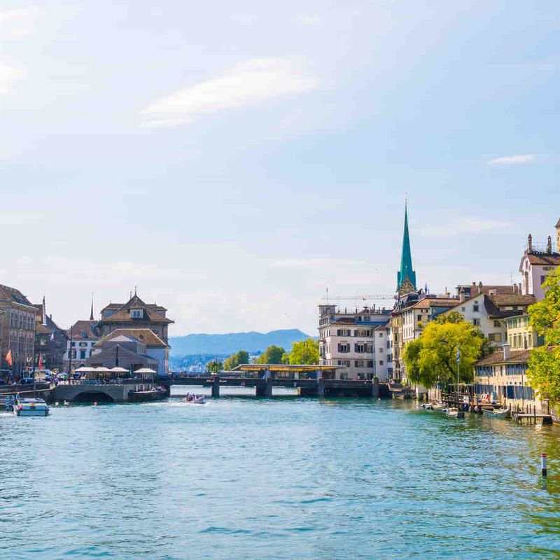 Panorama der Altstadt von Zürich mit dem Fluss Limmat, der zwischen historischen Gebäuden und Kirchtürmen fließt, an einem sonnigen Tag.
